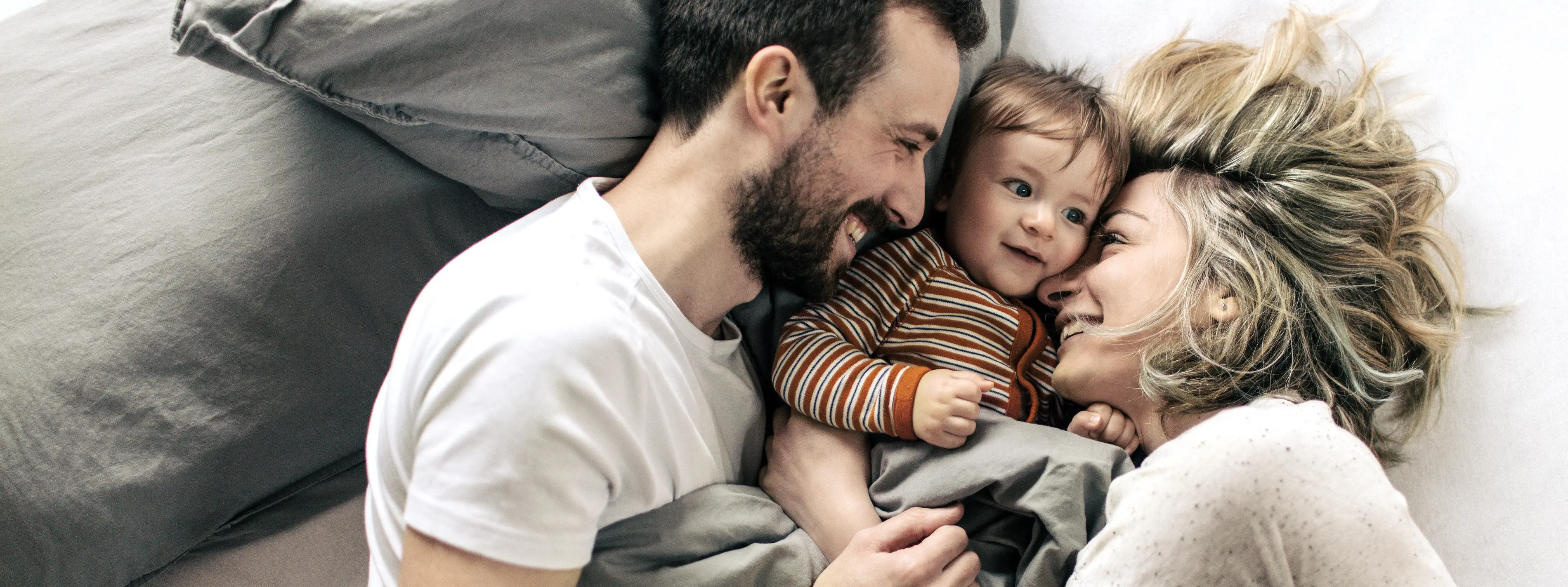 Eine Frau und ein Mann liegen sich zugewandt im Bett. Zwischen ihnen liegt ihr Kleinkind. Alle lächeln glücklich. | VIACTIV Krankenkasse