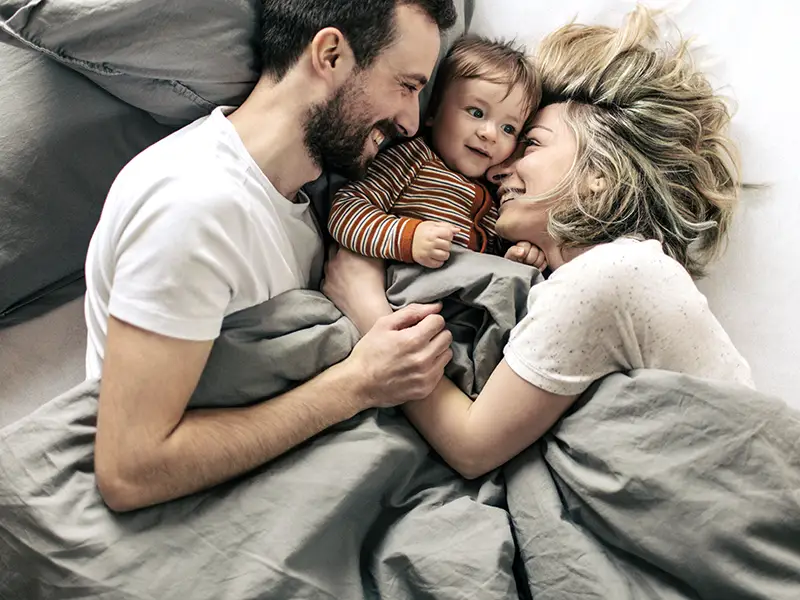 Eine Frau und ein Mann liegen sich zugewandt im Bett. Zwischen ihnen liegt ihr Kleinkind. Alle lächeln glücklich. | VIACTIV Krankenkasse