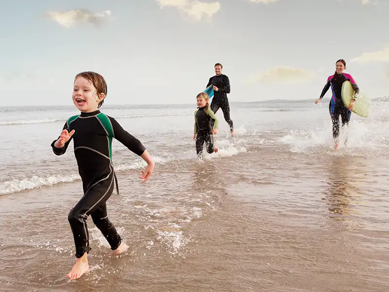 Eine vierköpfige Familie läuft in Neoprenanzügen am Strand entlang. | VIACTIV Krankenkasse