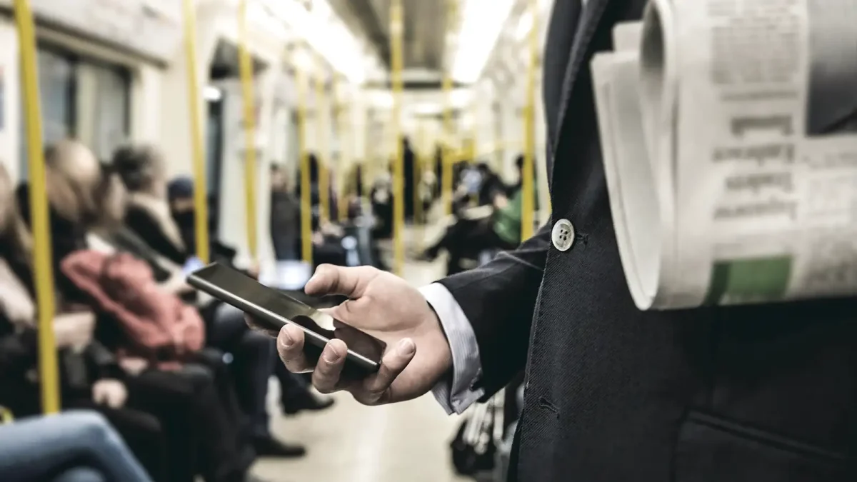 Ein Mann steht in einer vollen Straßenhahn. Er schaut auf sein Handy und hat eine Zeitung unter den Arm geklemmt. | VIACTIV Krankenkasse