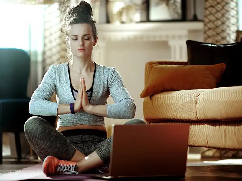 Eine junge Frau sitzt in einer Yogapose vor dem Laptop auf dem Boden in ihrem Wohnzimmer. Sie hat die Augen geschlossen. | VIACTIV Krankenkasse