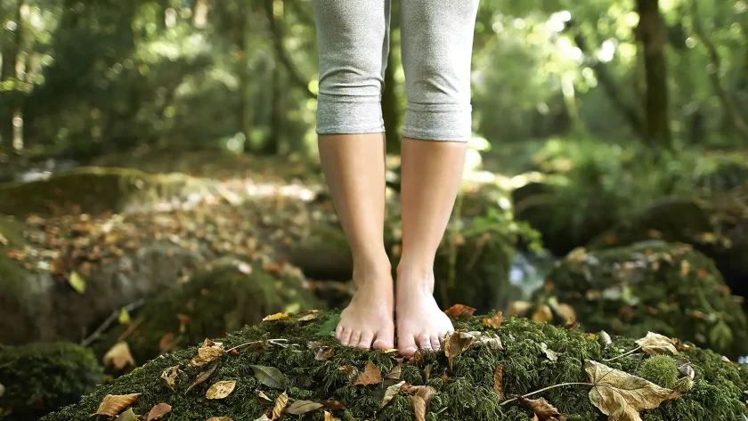 Eine Person steht mit nackten Füßen mitten im Wald bei Sonnenschein auf einem kleinen Hügel, der mit Moos bedeckt ist. | VIACTIV Krankenkasse