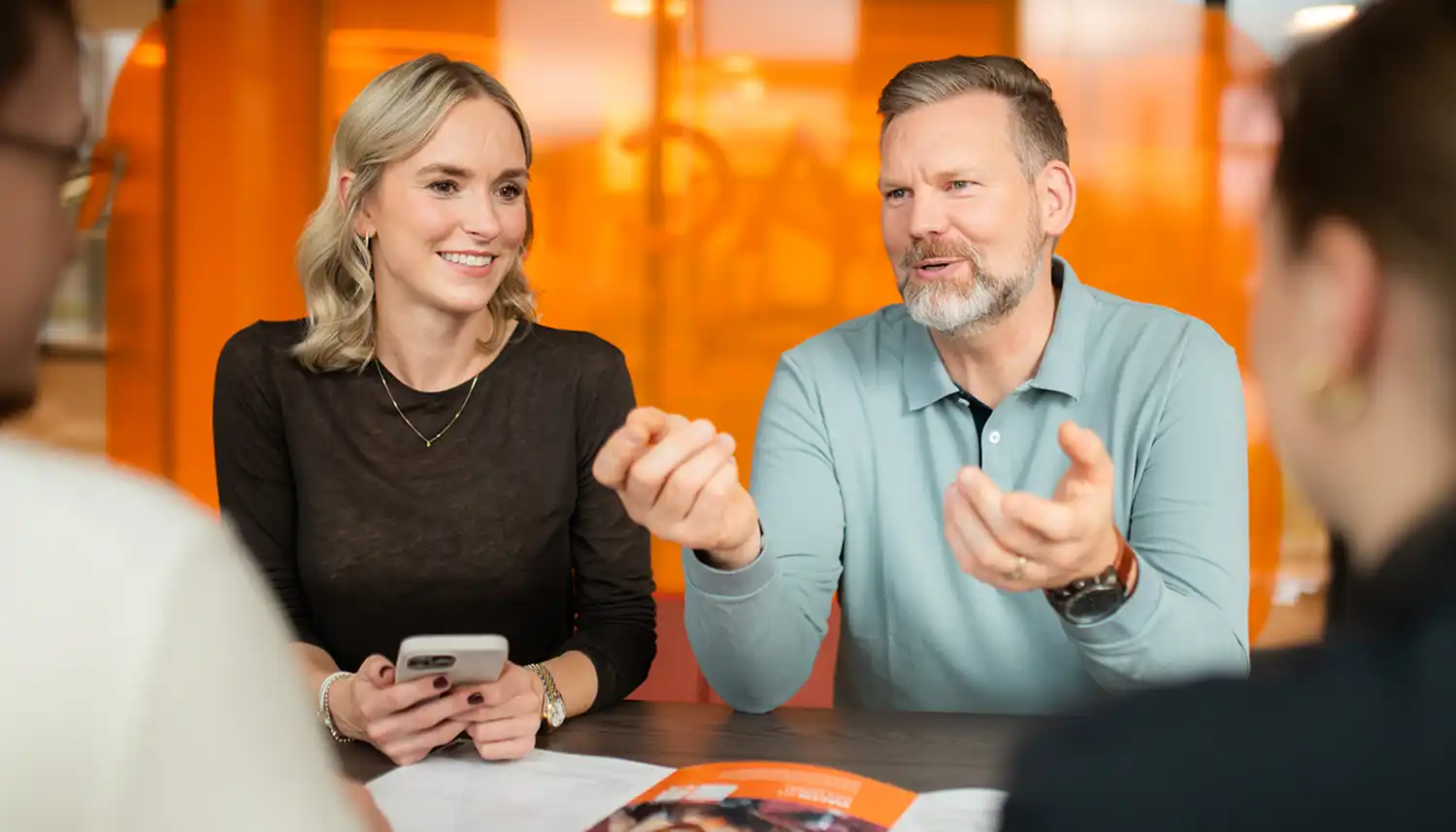 Eine Gruppe von VIACTIV Mitarbeitenden sitzt am Tisch. Sie unterhalten sich. Eine von ihnen hält ein Handy in der Hand. | VIACTIV Krankenkasse