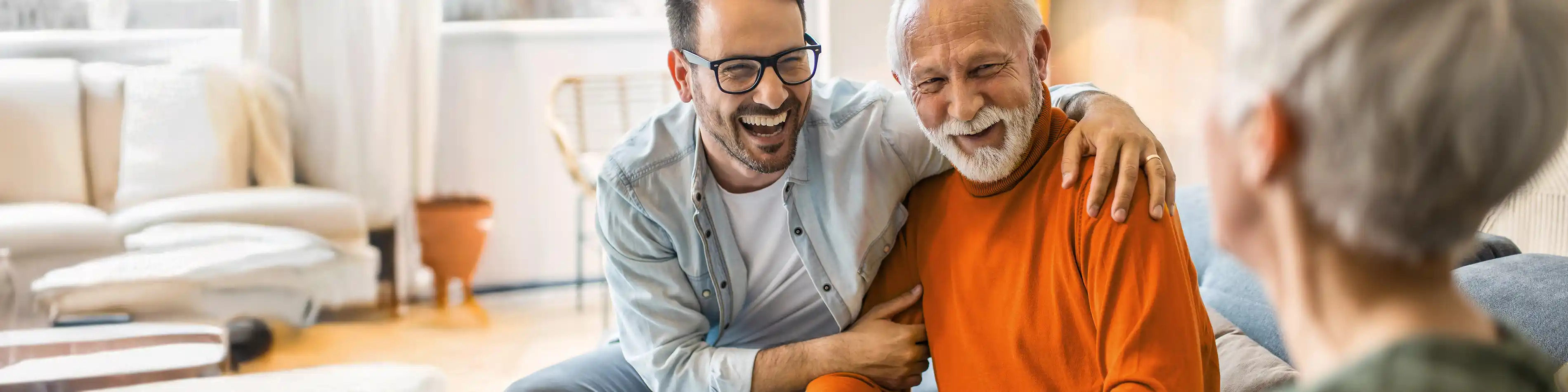 Ein jüngerer Mann umarmt einen älteren. Beide lachen und unterhalten sich mit einer Frau. | VIACTIV Krankenkasse