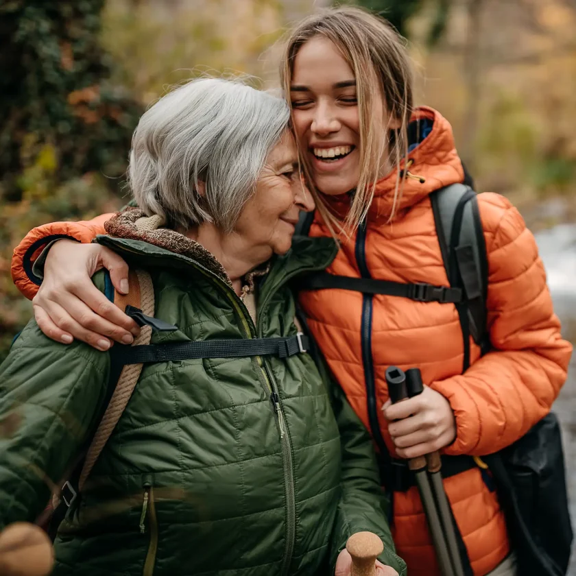 Eine jüngere und eine ältere Frau sind wandern in der Natur. Die jüngere Frau umarmt die Ältere. Beide lächeln. | VIACTIV Krankenkasse
