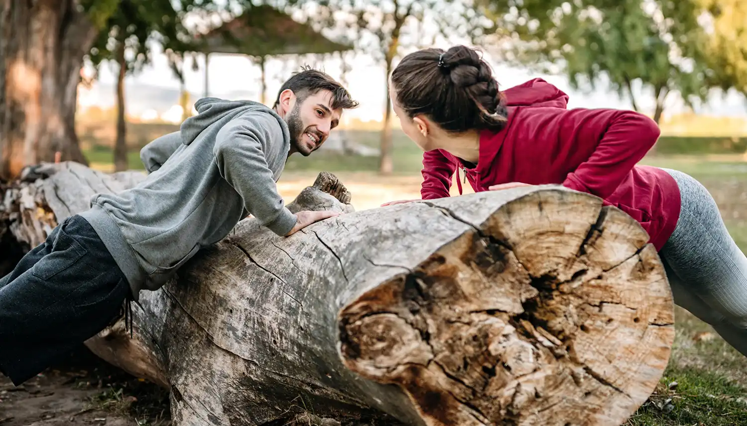 Junger Mann und junge Frau machen Outdoor Fitness an einem abgesägten Baum im Park bei Sonnenschein. | VIACTIV Krankenkasse