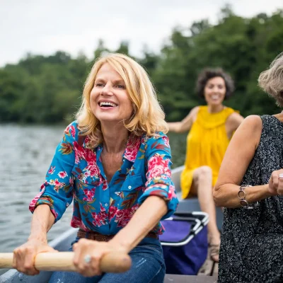 Drei Frauen sitzen an einem sonnigen Tag in einem Ruderboot und haben Spaß. Sie paddeln über einen See. Im Hintergrund sind Bäume. | VIACTIV Krankenkasse