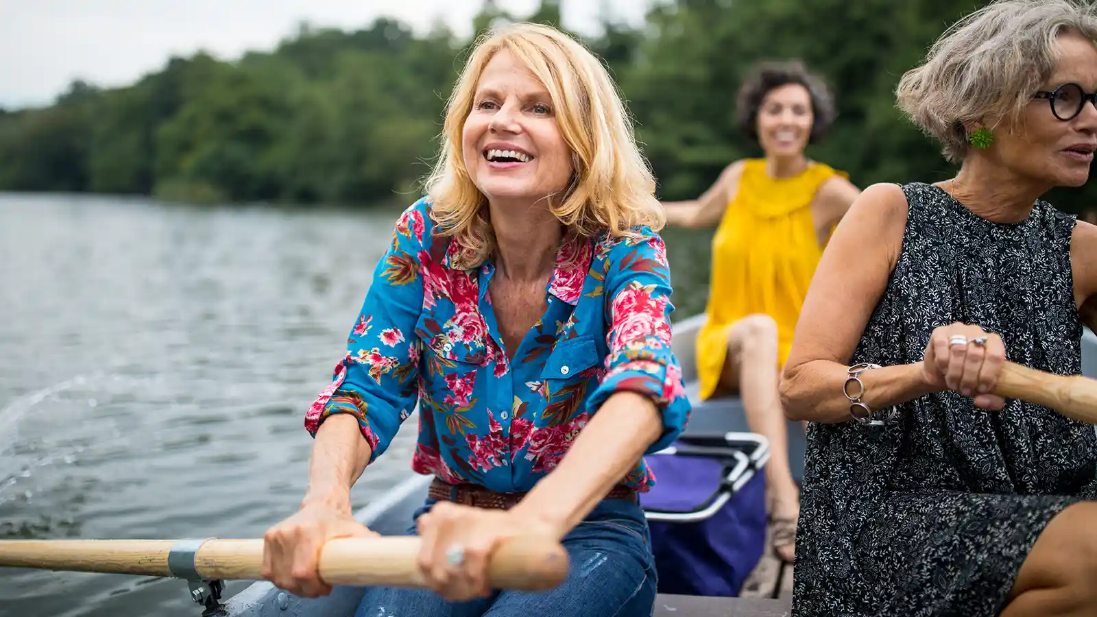 Drei Frauen sitzen an einem sonnigen Tag in einem Ruderboot und haben Spaß. Sie paddeln über einen See. Im Hintergrund sind Bäume. | VIACTIV Krankenkasse