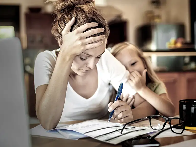 Eine Mutter sitzt am Schreibtisch. Sie hat ihren Kopf gestützt und wirkt gestresst. Hinter ihr ist ein Kind zu sehen. | VIACTIV Krankenkasse